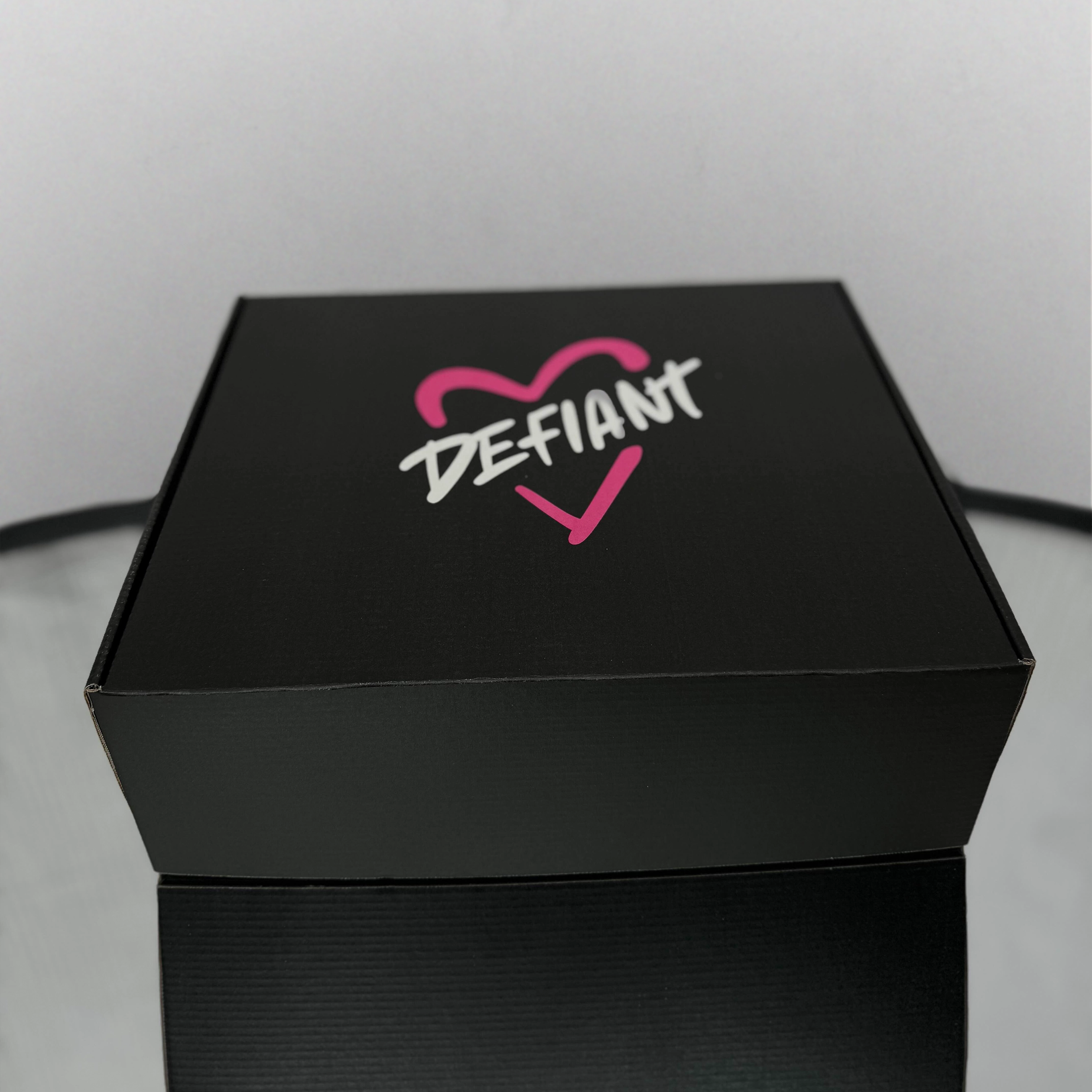 Black box with 'DEFIANT' logo featuring a pink heart on a light gray background